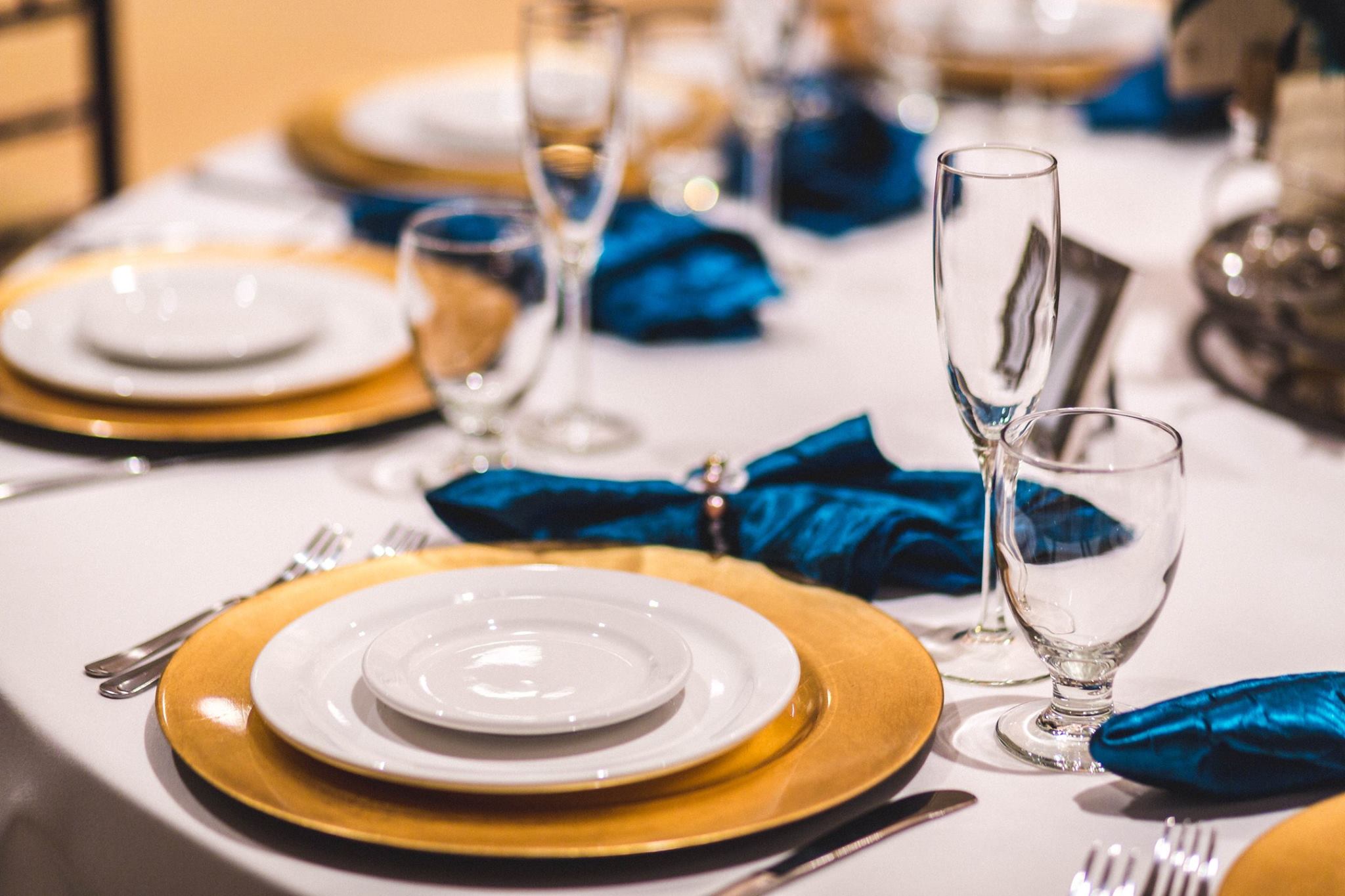 Showing a white tablecloth, white plates with brass accents, and blue napkins.